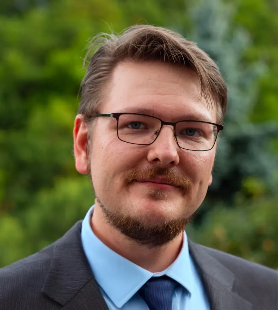 Portrait de Nicolas Nosal, lunettes, barbe courte, chemise bleue et veste sombre, devant un feuillage